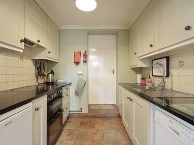 A galley kitchen with cream cabinets black countertops stove and sink at Manor Cottage in Northallerton