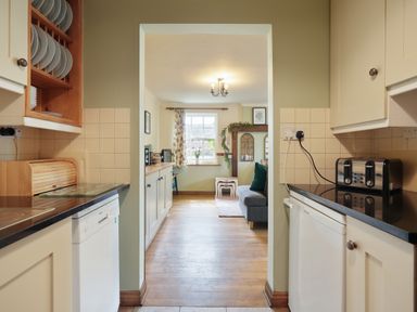 A kitchen with cabinets dishwasher and toaster looking into a living area with a window sofa and nesting tables at Manor Cottage in Northallerton
