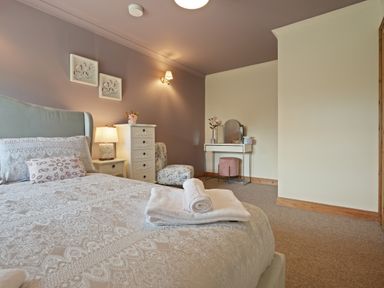 A bedroom with a bed white towels on the bed a nightstand with lamp a chest of drawers floral chair dressing table and stool at Manor Cottage in Northallerton