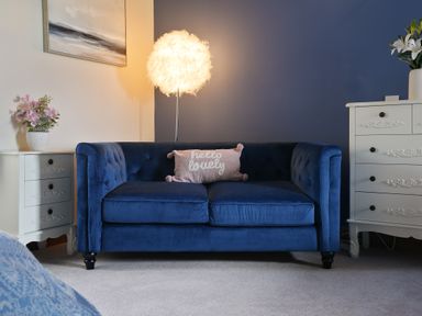 A blue sofa with a pillow in front of a blue wall with a standing lamp and white drawers with flowers at Manor Cottage in Northallerton