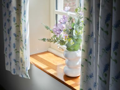 A window with floral curtains and a white vase with flowers on the wooden windowsill at Manor Cottage in Northallerton