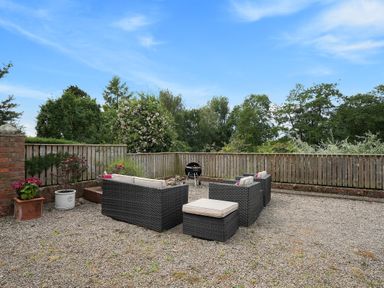 An outdoor seating area with rattan furniture and cushions on a gravel ground surrounded by wooden fencing and trees at Manor Cottage in Northallerton