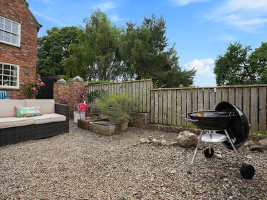 An outdoor patio area with a barbecue grill a cushioned sofa a pillow with hello summer text and a wooden fence at Manor Cottage in Northallerton
