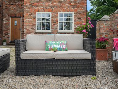 A patio with a black wicker sofa with beige cushions and a colorful pillow in front of a brick house with windows and a wooden door