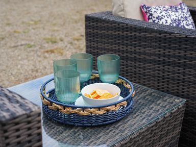 A wicker tray with four green glasses and a bowl of chips on a glass top outdoor table with wicker chairs at Manor Cottage in Northallerton