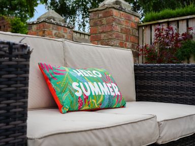An outdoor wicker sofa with beige cushions and a colorful pillow with hello summer text at Manor Cottage in Northallerton