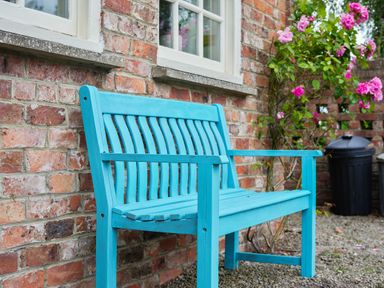 A blue wooden bench against a brick wall with a window above and pink flowers nearby at Manor Cottage in Northallerton