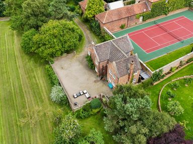 A brick house with a tiled roof next to a red and green tennis court and a gravel area with outdoor furniture surrounded by green lawns and trees at Manor Cottage in Northallerton