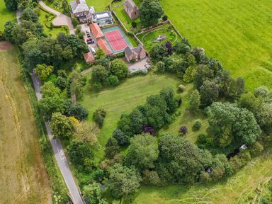 An aerial view of a property with buildings a tennis court gardens trees and grassy fields near a road at Manor Cottage in Northallerton