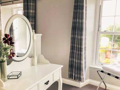 A white dressing table with a round mirror and flowers near a window with plaid curtains at Penlaurel in Launceston