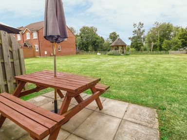 A wooden picnic table with a closed umbrella on a paved area facing a large grassy field with a gazebo and houses in the background at Ladybird in Bridgwater