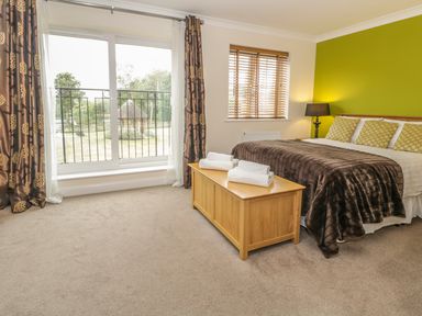 A bedroom with a double bed a wooden chest three pillows two folded towels a lamp window with blinds and sliding glass doors with curtains at Dragonfly in Bridgwater