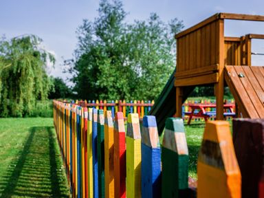A colorful wooden fence surrounding a green playground with a wooden play structure and picnic tables at Dragonfly in Bridgwater