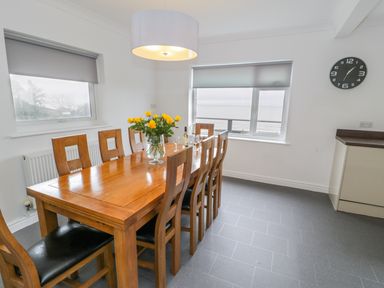 A dining area with a wooden table and chairs flowers on the table and windows showing an ocean view at Hendre Wylan in Llanaber