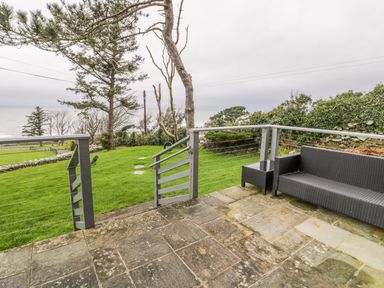 A patio with stone tiles and a wicker sofa overlooking a grassy yard with trees and an ocean view at Hendre Wylan in Llanaber