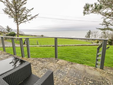 A patio with a wicker chair and a glass table overlooking a grassy field and the sea at Hendre Wylan in Llanaber