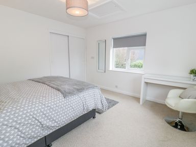 A bedroom with a bed covered in a patterned duvet next to a desk with a white chair and a window with a grey blind at Hendre Wylan in Llanaber