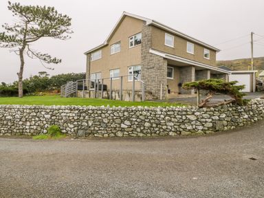 A two-story house with a stone wall in front and a tree on the lawn at Hendre Wylan in Llanaber