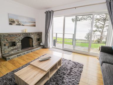 A living room with a stone fireplace a wooden coffee table gray curtains and large sliding glass doors overlooking a garden and trees at Hendre Wylan in Llanaber