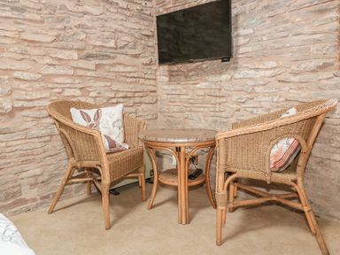 A corner seating area with two wicker chairs with cushions and a small round table under a wall-mounted TV at The Cow Cott in Hereford