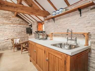 A kitchen with wooden cabinets, a stainless steel sink, a toaster, a shelf with containers and two wicker chairs at The Cow Cott in Hereford