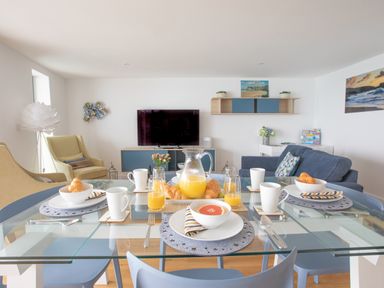 A dining table set with breakfast items in a living room with chairs a television and wall shelves at Onshore 6 - Newquay Towan Beach View in Newquay