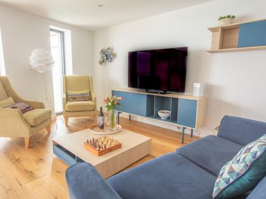 A living room with armchairs a sofa a coffee table with a chessboard flowers and wine a television on a cabinet and a floor lamp at Onshore 6 - Newquay Towan Beach View in Newquay