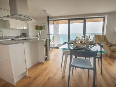 A kitchen and dining area with glass table set for breakfast and open doors to a balcony overlooking the sea at Onshore 6 - Newquay Towan Beach View in Newquay