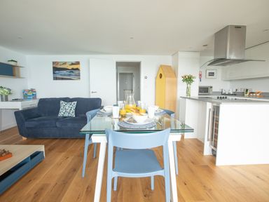 A dining area with a glass table set for breakfast and blue chairs next to a living room with a blue sofa and kitchen with white cabinets at Onshore 6 - Newquay Towan Beach View in Newquay