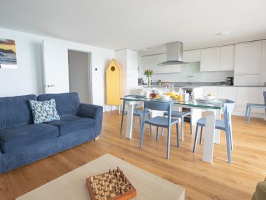 An open living space with a blue sofa a glass dining table with blue chairs and a kitchen with white cabinets at Onshore 6 - Newquay Towan Beach View in Newquay