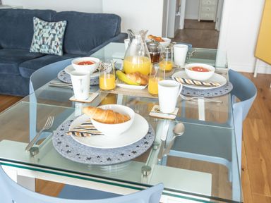 A glass dining table set for breakfast with croissants, grapefruit bowls, orange juice, and mugs in a room with a blue sofa and wooden floor at Onshore 6 - Newquay Towan Beach View in Newquay