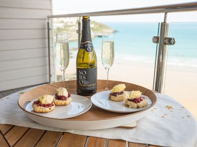 A wooden table on a balcony with a bottle of prosecco two filled glasses and two plates with scones and cream with a beach view at Onshore 6 - Newquay Towan Beach View in Newquay