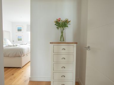 A chest of drawers with a vase of flowers on top in a hallway next to a door and a view of a bedroom with a bed and picture frames at Onshore 6 - Newquay Towan Beach View in Newquay