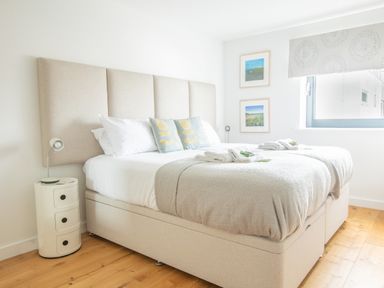 A bedroom with a bed with two blue pillows and white linens beside a round white bedside table with a lamp at Onshore 6 - Newquay Towan Beach View in Newquay
