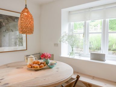 A dining area with a round wooden table set with pastries teapot and flowers near a window at Whitehall Farm Cottage in Honiton