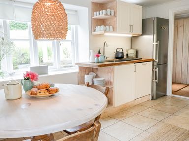 A kitchen with a round wooden table with pastries and a teapot next to a window and a counter with a sink and shelves holding dishes at Whitehall Farm Cottage in Honiton