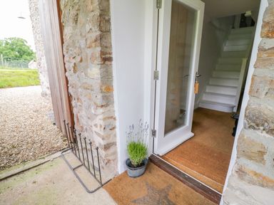 An entrance with a glass door opening to a hallway with white stairs and a doormat outside the door at Whitehall Farm Cottage in Honiton