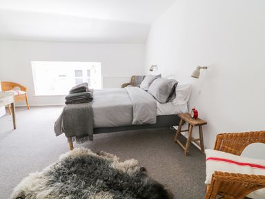 A bedroom with a bed with grey bedding two wicker chairs a wooden stool with a red alarm clock and a sheepskin rug on the floor at Whitehall Farm Cottage in Honiton