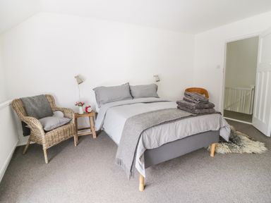 A bedroom with a double bed and gray bedding a wicker chair with a gray blanket and pillow and a wooden side table with flowers and a clock at Whitehall Farm Cottage in Honiton