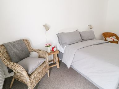A bedroom with a bed with gray bedding wicker chairs and a wooden side table with flowers and an alarm clock at Whitehall Farm Cottage in Honiton