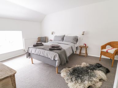 A bedroom with a double bed with gray bedding two wicker chairs a wooden stool with a red alarm clock and a fur rug on the floor at Whitehall Farm Cottage in Honiton