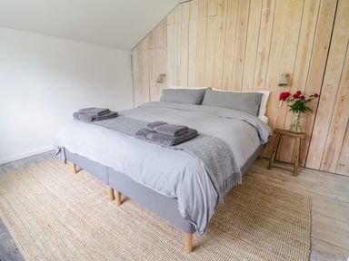 A bedroom with a bed covered in gray bedding and towels with a wooden stool holding a vase of flowers at Whitehall Farm Cottage in Honiton