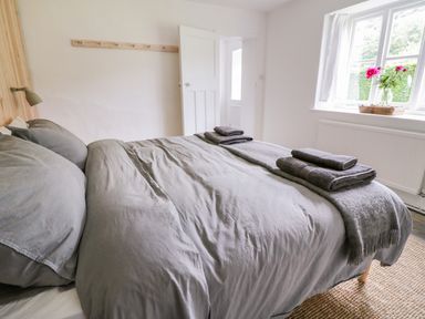 A bedroom with a bed made with gray linens and folded towels a window with flowers and an open door at Whitehall Farm Cottage in Honiton