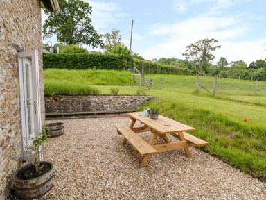 A gravel patio with a wooden picnic table and potted plants next to a stone building with a door open to a grassy field at Whitehall Farm Cottage in Honiton