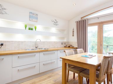 A kitchen with a wooden dining table and wicker chairs near large windows at 6 Lake View in Lanreath