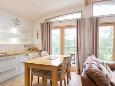 A kitchen dining area with wooden table and chairs next to glass doors and a brown leather sofa at 6 Lake View in Lanreath