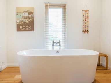 A bathroom with a freestanding bathtub a window with blinds a wooden stool a wall hanging of shells and a Rock Cornwall poster at 6 Lake View in Lanreath