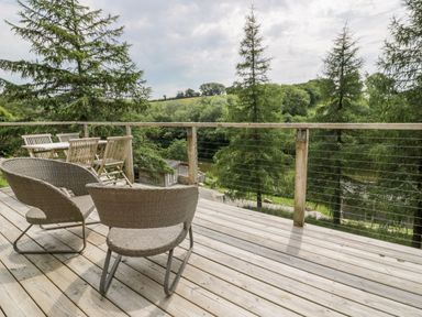 An outdoor wooden deck with wicker chairs and a table surrounded by trees at 6 Lake View in Lanreath