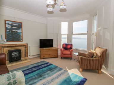A living room with armchairs a television on a wooden cabinet a fireplace and a large window overlooking the sea at 11 Marine Terrace in Criccieth