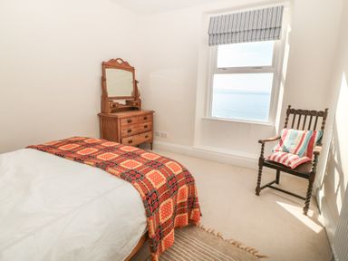 A bedroom with a bed covered with a patterned blanket a wooden dresser with a mirror wooden chair with striped cushions by a window showing a sea view at 11 Marine Terrace in Criccieth
